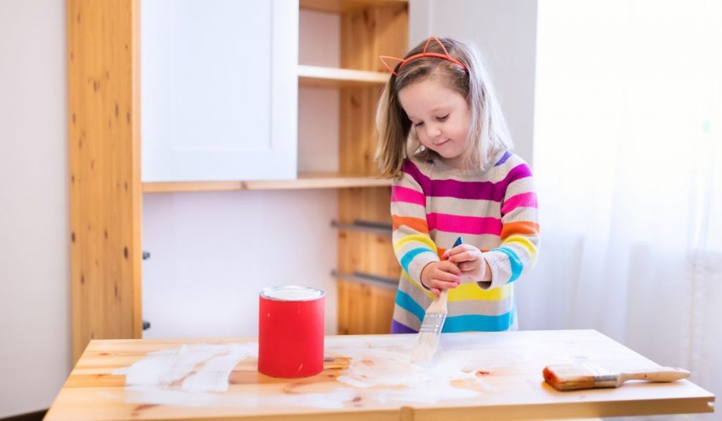 a preschool girl holding a brush painting the wooden table with white paint